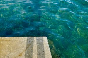 A concrete slab with shadows from a pair of legs, blue green water below. 