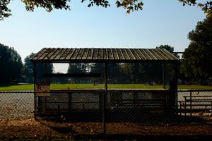 Metal-roofed dugout between a green baseball field and a chain link fence, bleachers to the right, blue and white sky above.