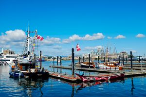 Inner harbour in Victoria, BC. A few boats are docked, two Canadian flags are flying. Blue sky with a few clouds.